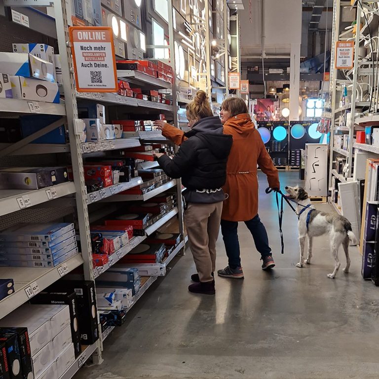 Zwei Personen und ein Hund in einem Baumarkt, schauen sich Regale mit Produkten an. Übung Hundeführerschein