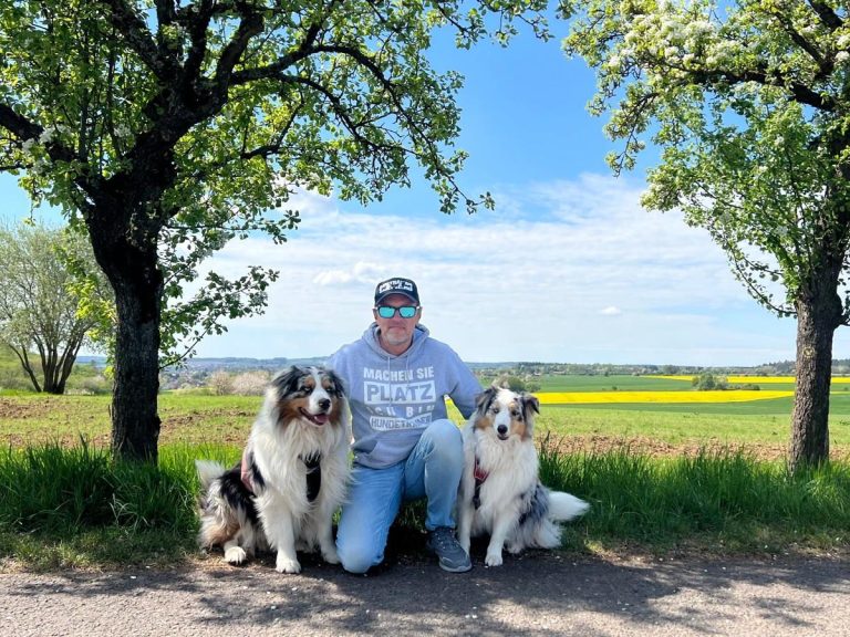 Michael Szagarus Hundetrainer mit Bao und Bandhu Person sitzt zwischen zwei Hunden unter Bäumen auf einem gepflasterten Weg.