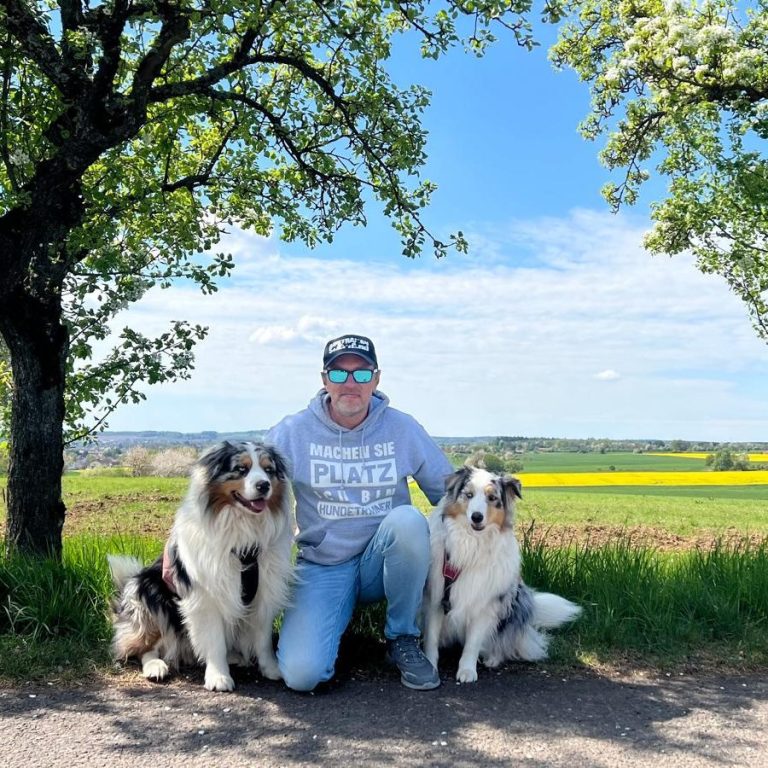 Hundetrainer Michael Szagarus Mann sitzt auf dem Boden zwischen zwei Hunden unter einem Baum, vor einer blühenden Landschaft.